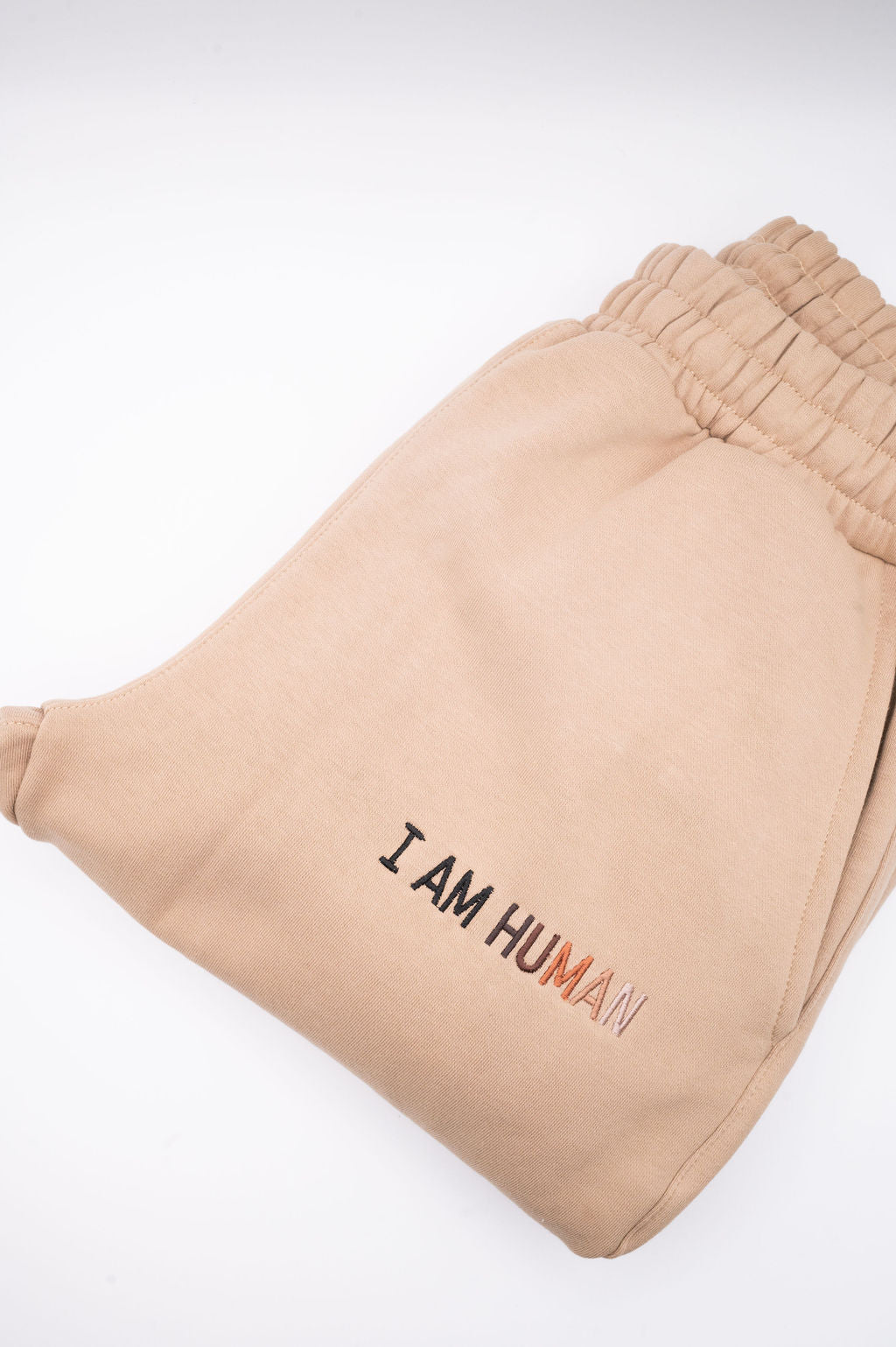 A folded pair of tan I AM HUMAN logo joggers showing the stretch waistband and embroidered logo. 